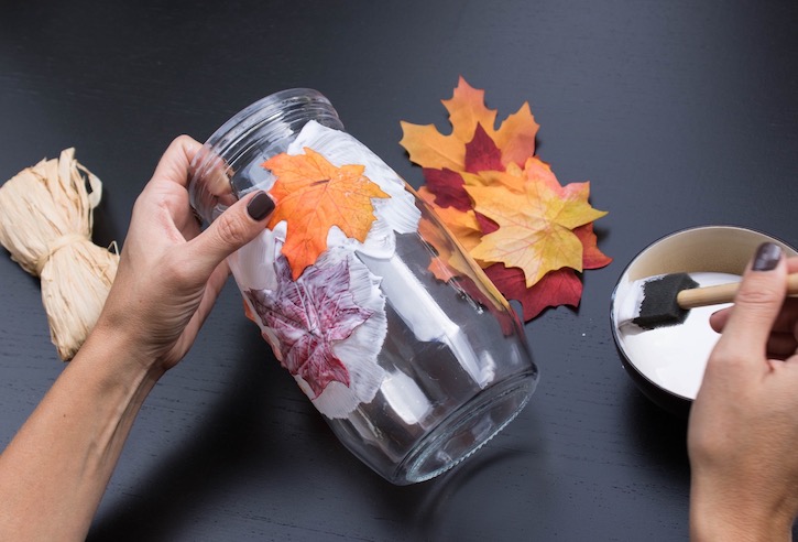 Using a sponge brush and mod podge to apply faux leaves to a jar to create a stunning DIY Fall Leaf Candle Holder. 