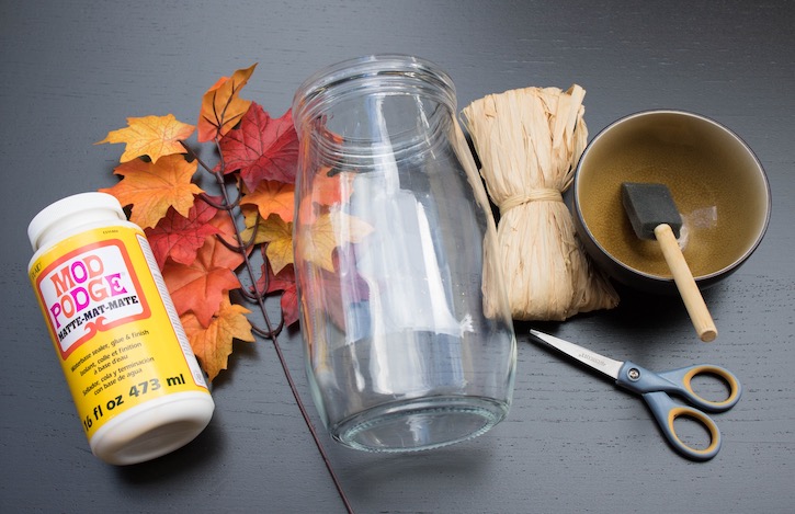 Supplies needed to make a DIY Fall Leaf Candle Holder: Any glass jar, matte mod podge, fall leaves, a spronge brush, and raffia for the finishing touch. 