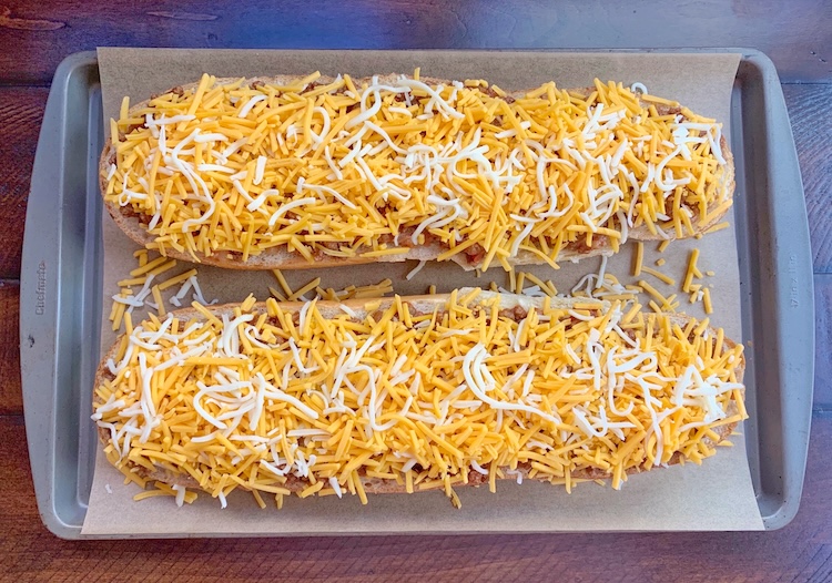 French bread sliced in half on a baking sheet topped with Sloppy Joe Mix and lots of shredded cheese, ready to go into the oven and bake. 