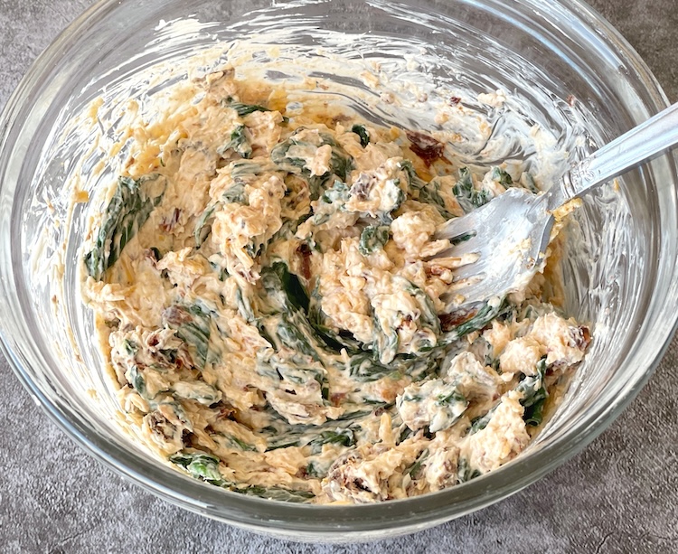 A mixture of cream cheese, cheddar cheese, sun-dried tomatoes, spinach, and garlic powder in a mixing bowl getting ready to top chicken breasts for baking a low carb meal. 