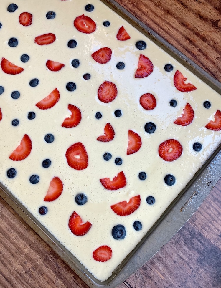 Pancake batter on a sheet pan topped with fresh sliced strawberries and blueberries ready for the oven to make breakfast for a crowd. 