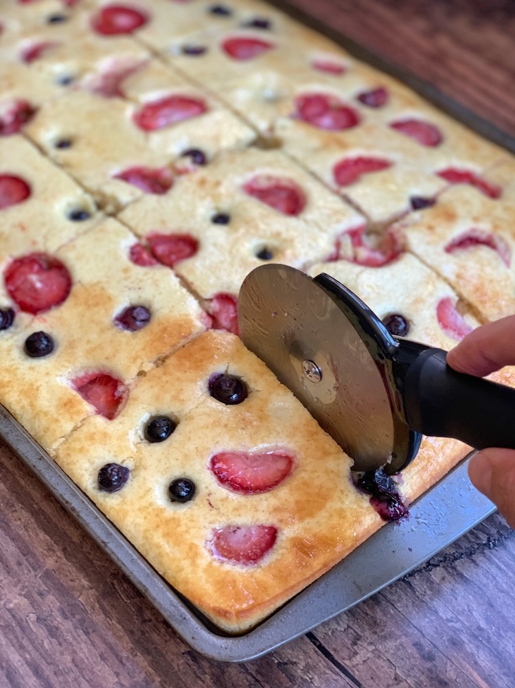 Baked pancakes on a sheet pan being cut with a pizza cutter to make individual slices to feed a crowd at breakfast time.
